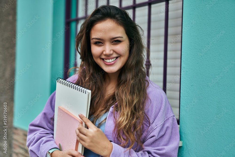 Fototapeta premium Young hispanic student woman smiling happy holding books at the city.