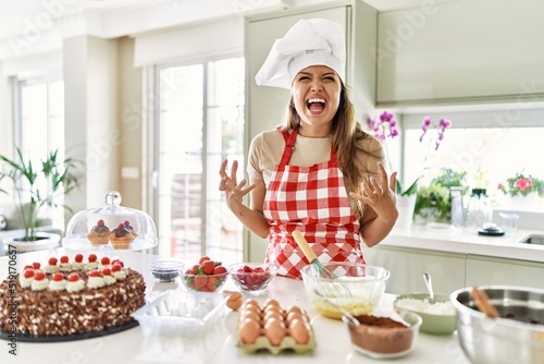 Beautiful young brunette pastry chef woman cooking pastries at the kitchen crazy and mad shouting and yelling with aggressive expression and arms raised. frustration concept.