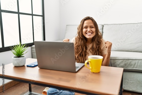 Beautiful hispanic woman using computer laptop at home screaming proud, celebrating victory and success very excited with raised arms