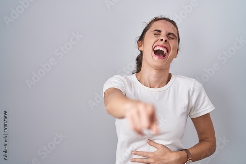 Beautiful brunette woman standing over isolated background laughing at you, pointing finger to the camera with hand over body, shame expression