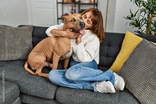 Young caucasian woman kissing and hugging dog sitting on sofa at home