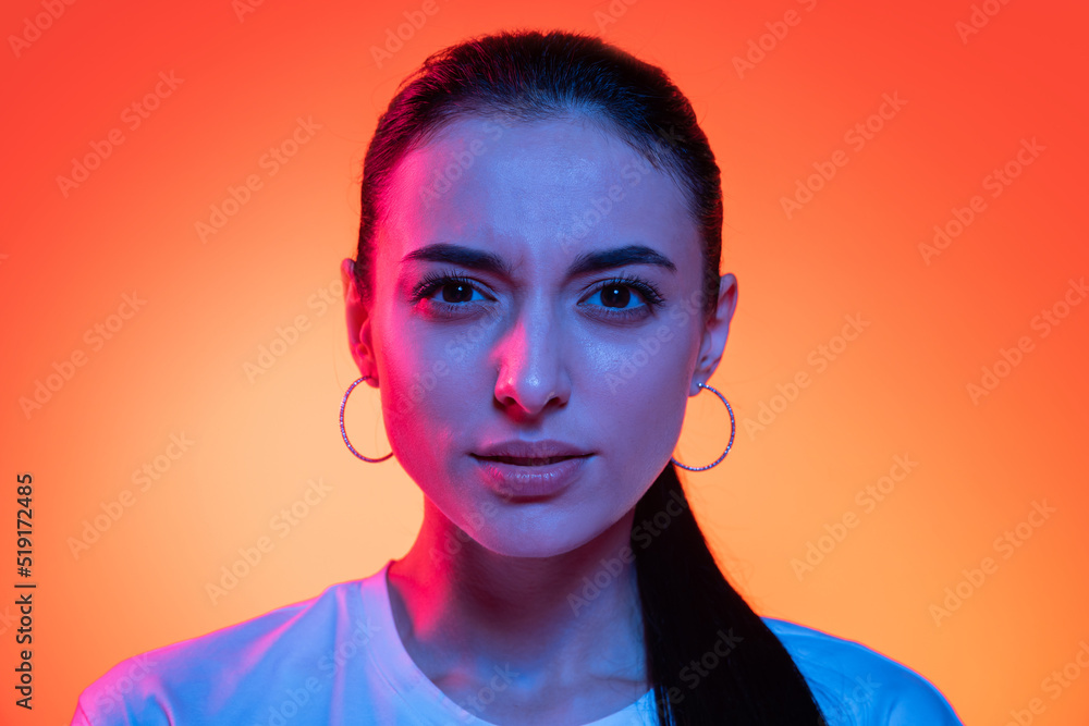 Closeup face of young attractive girl looking at camera isolated on orange color background in neon light. Concept of emotions, facial expressions Closeup face of young attractive girl looking at camera isolated on orange color background in neon light. Concept of emotions, facial expressions