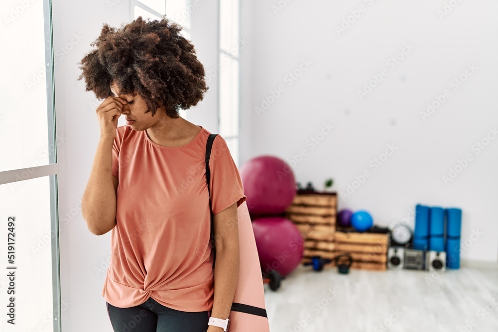 African american woman with afro hair holding yoga mat at pilates room tired rubbing nose and eyes feeling fatigue and headache. stress and frustration concept. African american woman with afro hair holding yoga mat at pilates room tired rubbing nose and eyes feeling fatigue and headache. stress and frustration concept.