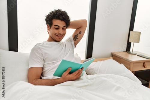 Young hispanic man reading book sitting on the bed at bedroom.