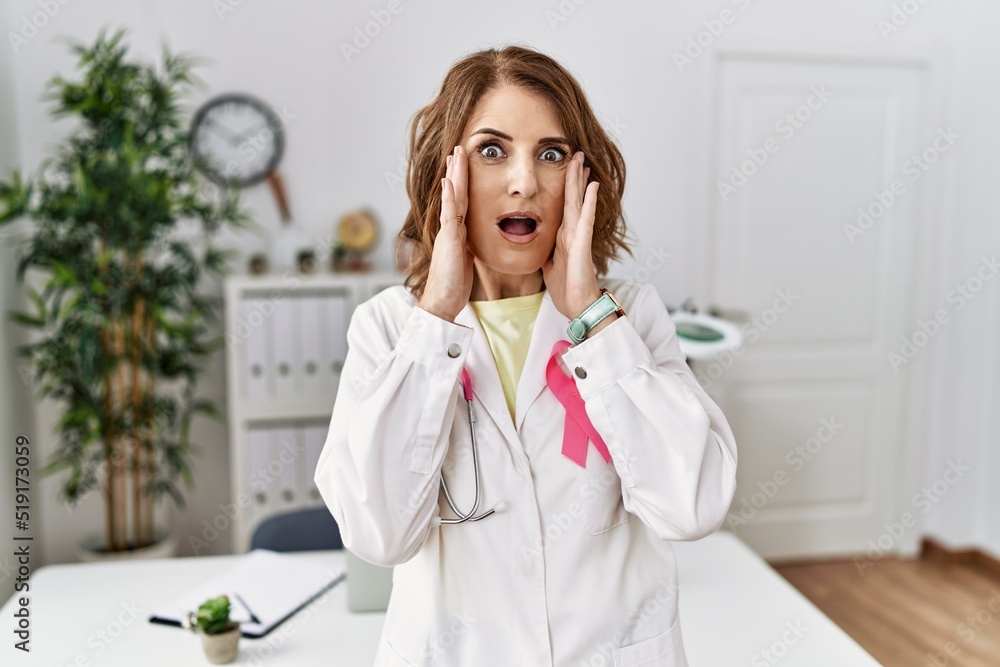 Middle age doctor woman wearing pink cancer ribbon on uniform afraid and shocked, surprise and amazed expression with hands on face Middle age doctor woman wearing pink cancer ribbon on uniform afraid and shocked, surprise and amazed expression with hands on face