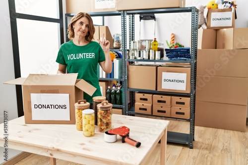 Middle age woman wearing volunteer t shirt at donations stand surprised with an idea or question pointing finger with happy face, number one © Krakenimages.com