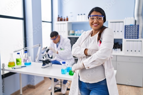 Young hispanic woman expecting a baby working at scientist laboratory happy face smiling with crossed arms looking at the camera. positive person.