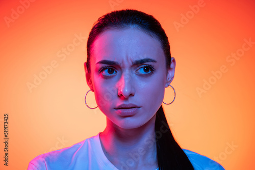 Closeup face of young attractive girl looking at camera isolated on orange color background in neon light. Concept of emotions  facial expressions