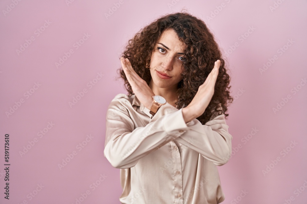 Hispanic woman with curly hair standing over pink background rejection expression crossing arms doing negative sign, angry face Hispanic woman with curly hair standing over pink background rejection expression crossing arms doing negative sign, angry face