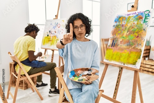 Young hispanic woman at art studio pointing with finger up and angry expression, showing no gesture