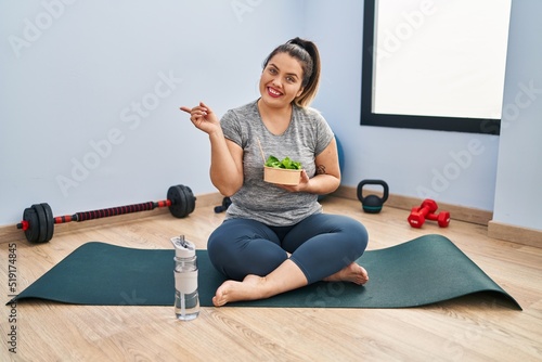 Young hispanic woman sitting on yoga mat eating healthy salad smiling happy pointing with hand and finger to the side