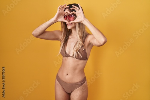 Young hispanic woman wearing bikini over yellow background doing heart shape with hand and fingers smiling looking through sign