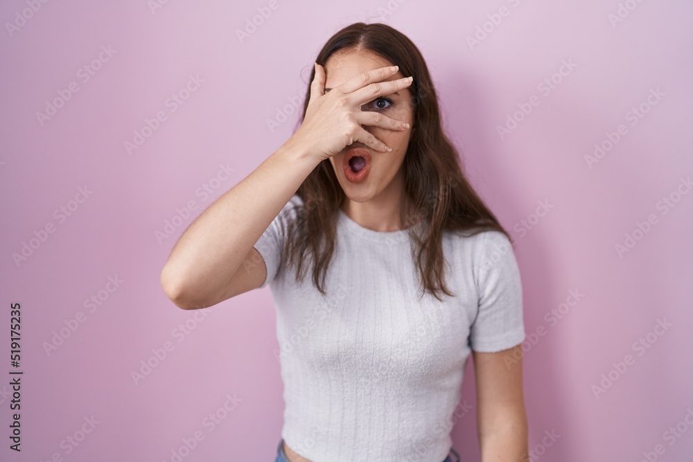 Young hispanic girl standing over pink background peeking in shock covering face and eyes with hand, looking through fingers with embarrassed expression. Young hispanic girl standing over pink background peeking in shock covering face and eyes with hand, looking through fingers with embarrassed expression.