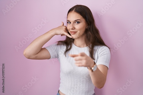 Young hispanic girl standing over pink background smiling doing talking on the telephone gesture and pointing to you. call me.