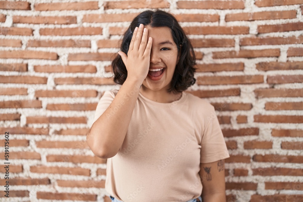 Young hispanic woman standing over bricks wall covering one eye with hand, confident smile on face and surprise emotion. Young hispanic woman standing over bricks wall covering one eye with hand, confident smile on face and surprise emotion.
