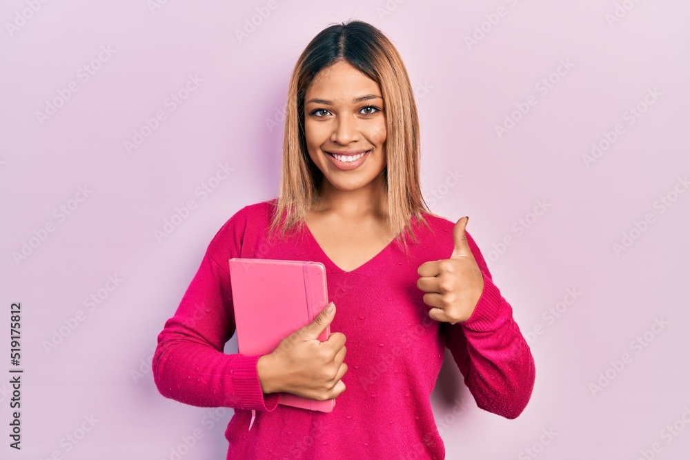 Beautiful hispanic woman holding book smiling happy and positive, thumb up doing excellent and approval sign Beautiful hispanic woman holding book smiling happy and positive, thumb up doing excellent and approval sign