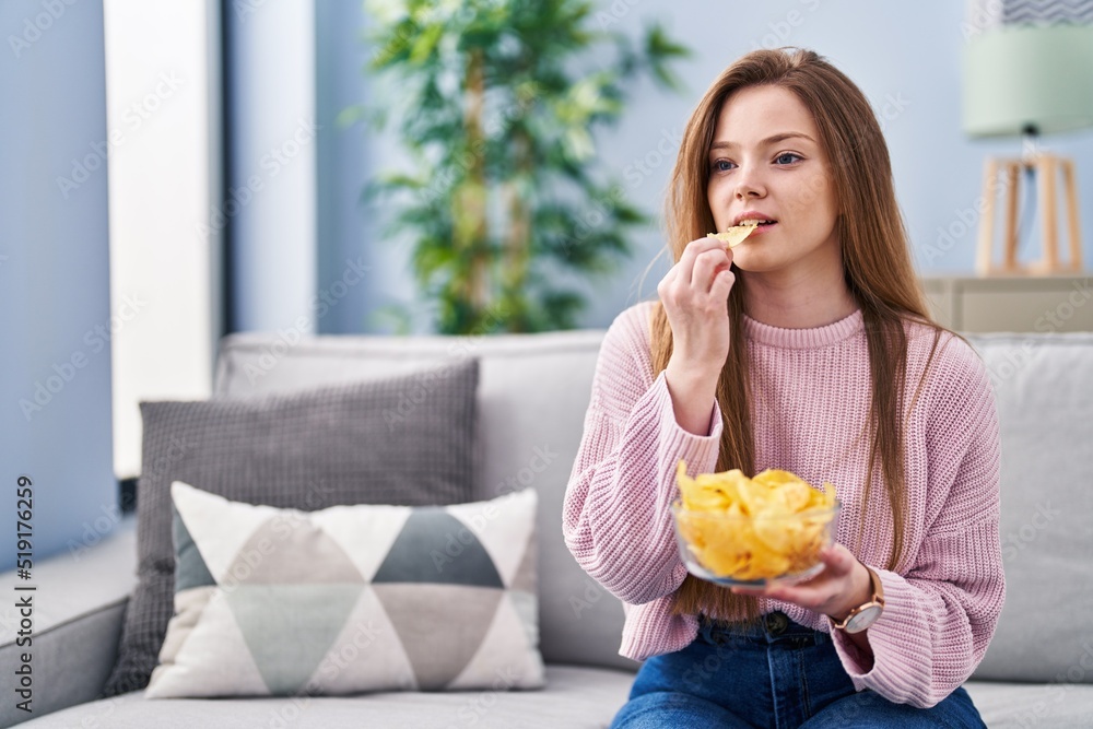 Young caucasian woman eating chips potatoes sitting on sofa at home Young caucasian woman eating chips potatoes sitting on sofa at home