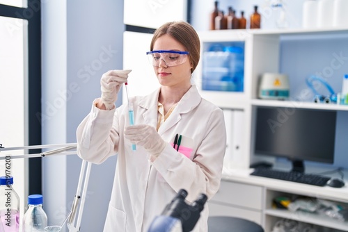 Young blonde woman wearing scientist uniform measuring liquid on test tube at laboratory