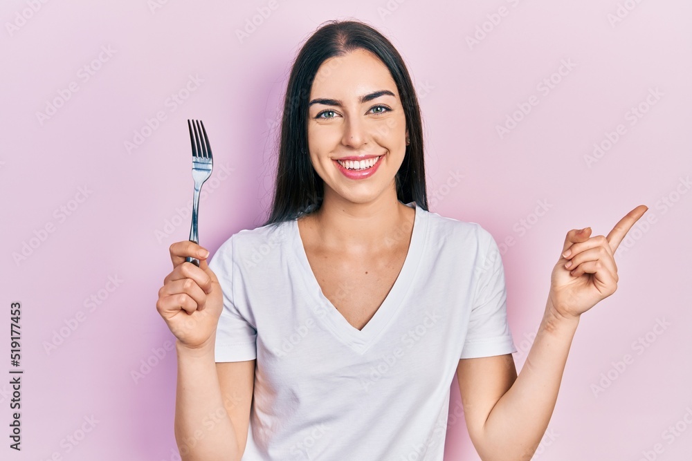 Beautiful woman with blue eyes holding one silver fork smiling happy pointing with hand and finger to the side Beautiful woman with blue eyes holding one silver fork smiling happy pointing with hand and finger to the side