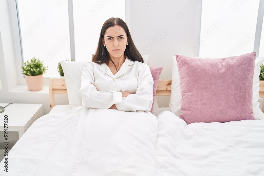 Young hispanic woman sitting on bed with arms crossed gesture and serious expression at bedroom