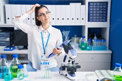 Young brunette woman working at scientist laboratory confuse and wondering about question. uncertain with doubt  thinking with hand on head. pensive concept.