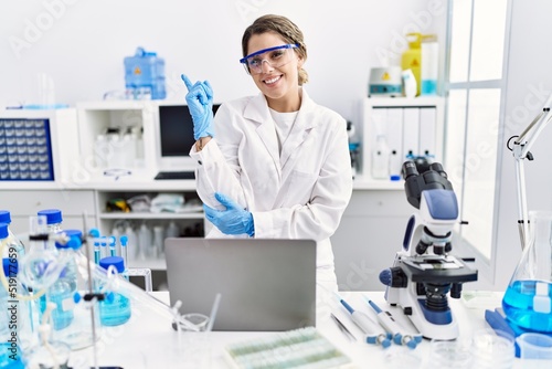 Young hispanic woman wearing scientist uniform working at laboratory smiling happy pointing with hand and finger to the side
