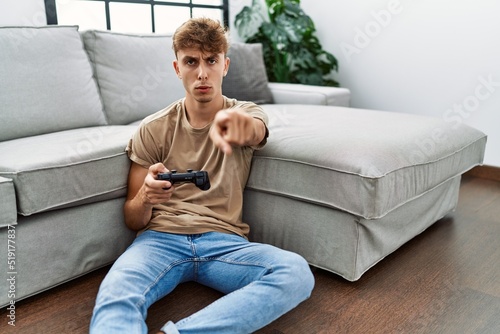 Young caucasian man playing video game holding controller at home pointing with finger to the camera and to you, confident gesture looking serious