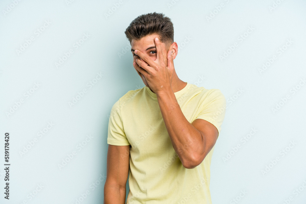 Young caucasian man isolated on blue background blink at the camera through fingers, embarrassed covering face.