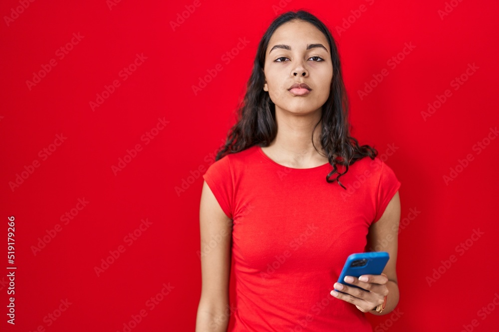 Young brazilian woman using smartphone over red background relaxed with serious expression on face. simple and natural looking at the camera. Young brazilian woman using smartphone over red background relaxed with serious expression on face. simple and natural looking at the camera.