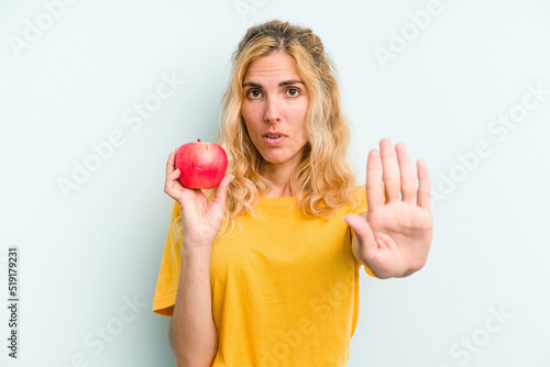 Young caucasian woman holding an apple isolated on blue background standing with outstretched hand showing stop sign, preventing you.