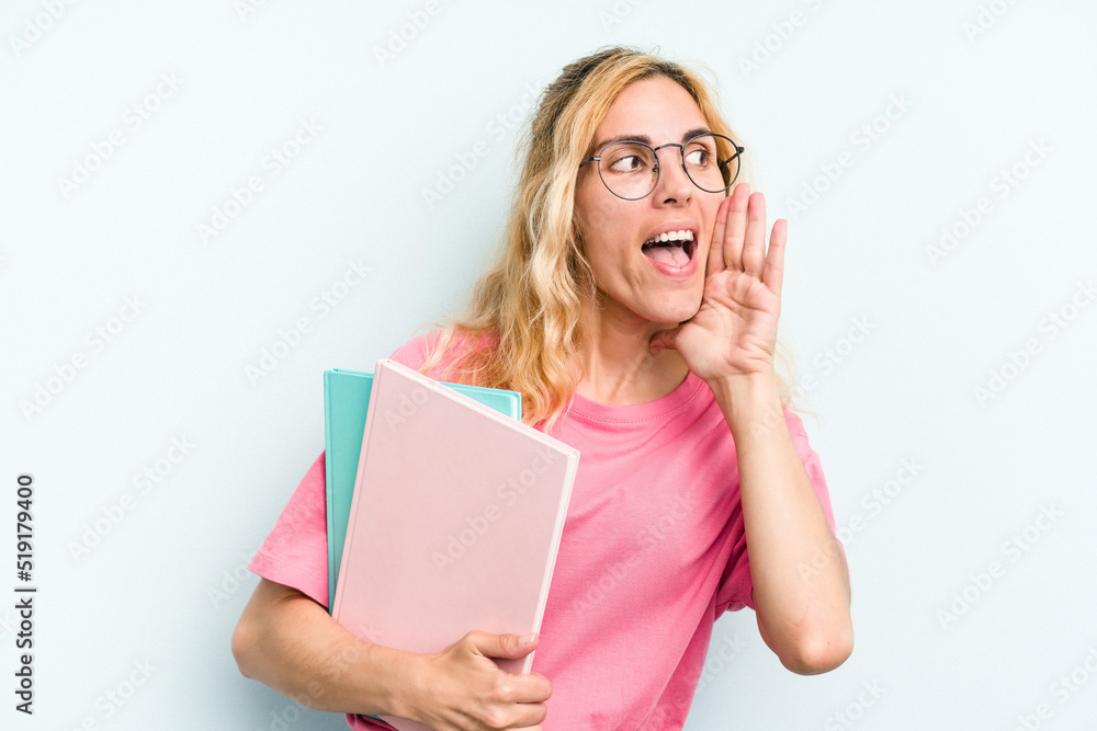 Young student caucasian woman holding books isolated on blue background shouting and holding palm near opened mouth. Young student caucasian woman holding books isolated on blue background shouting and holding palm near opened mouth.