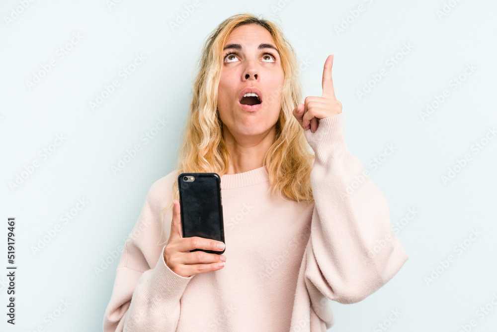 Young caucasian woman holding mobile phone isolated on blue background pointing upside with opened mouth. Young caucasian woman holding mobile phone isolated on blue background pointing upside with opened mouth.