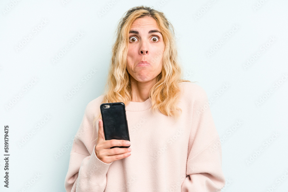 Young caucasian woman holding mobile phone isolated on blue background shrugs shoulders and open eyes confused. Young caucasian woman holding mobile phone isolated on blue background shrugs shoulders and open eyes confused.