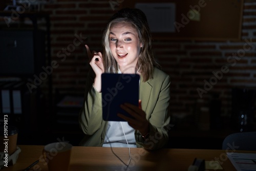 Blonde caucasian woman working at the office at night with a big smile on face, pointing with hand finger to the side looking at the camera.