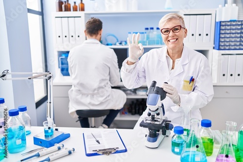 Middle age woman working at scientist laboratory doing ok sign with fingers  smiling friendly gesturing excellent symbol