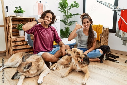 Photography Young hispanic couple doing laundry with dogs smiling doing phone gesture with hand and fingers like talking on the telephone