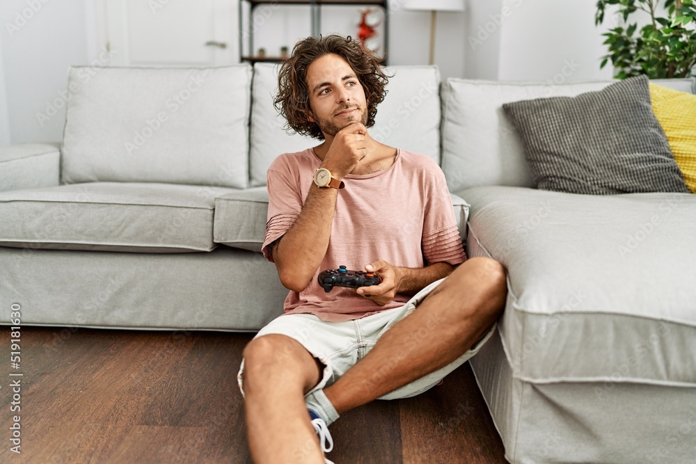 Obraz premium Young hispanic man playing video game holding controller at home serious face thinking about question with hand on chin, thoughtful about confusing idea