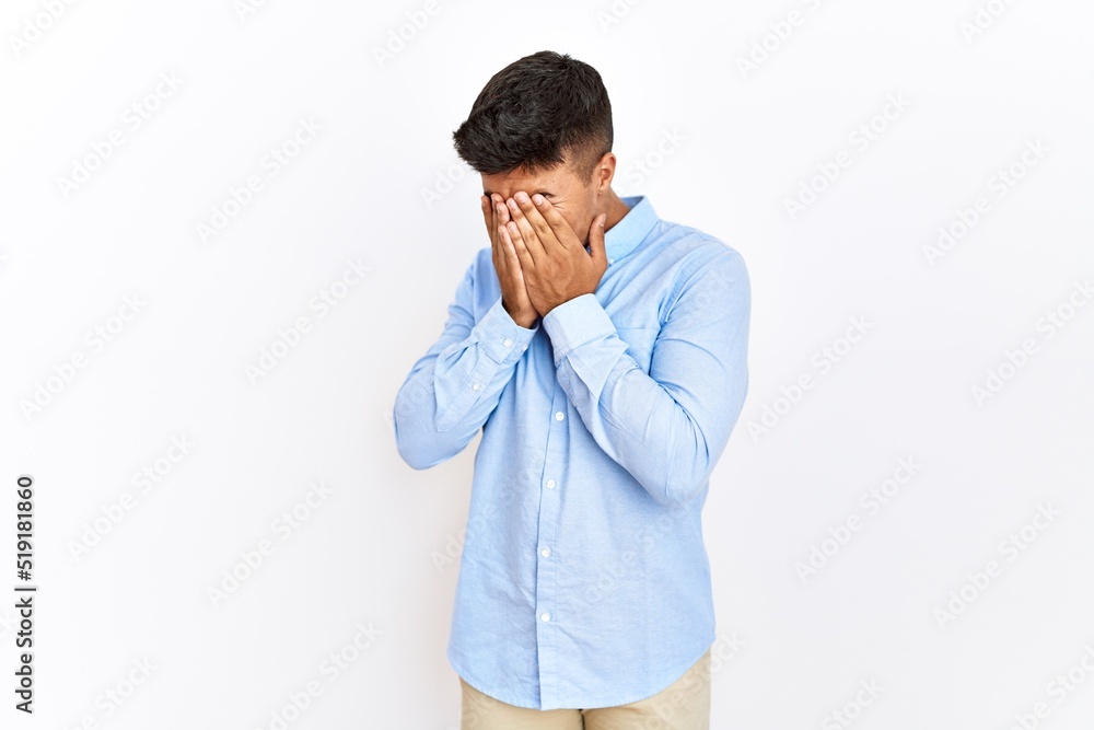 Young hispanic man wearing business shirt standing over isolated background with sad expression covering face with hands while crying. depression concept. Young hispanic man wearing business shirt standing over isolated background with sad expression covering face with hands while crying. depression concept.