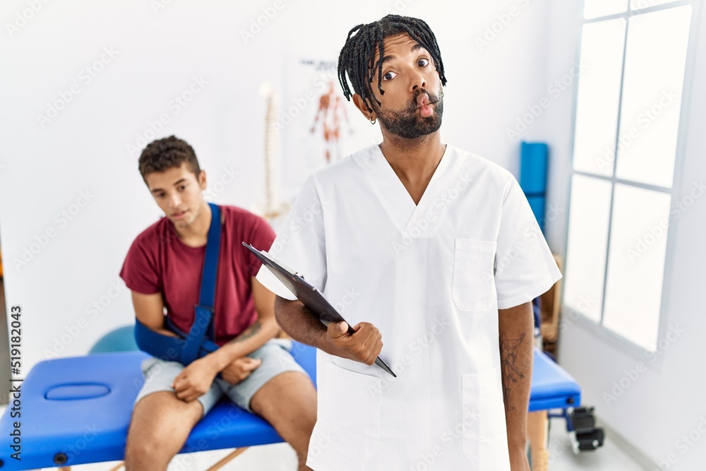 Obraz premium Young hispanic man working at pain recovery clinic with a man with broken arm making fish face with lips, crazy and comical gesture. funny expression.