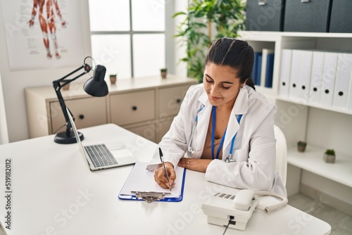 Young hispanic woman wearing doctor uniform writing medical report at clinic