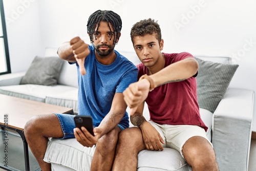 Young hispanic men using smartphone sitting on the sofa at home looking unhappy and angry showing rejection and negative with thumbs down gesture. bad expression.