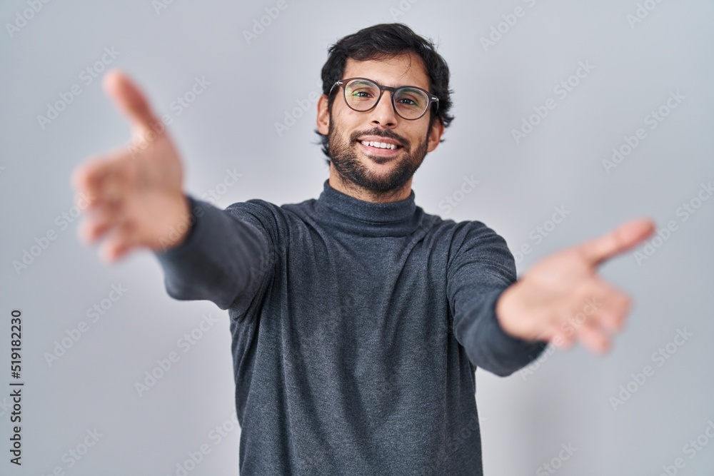 Handsome latin man standing over isolated background looking at the camera smiling with open arms for hug. cheerful expression embracing happiness. Handsome latin man standing over isolated background looking at the camera smiling with open arms for hug. cheerful expression embracing happiness.