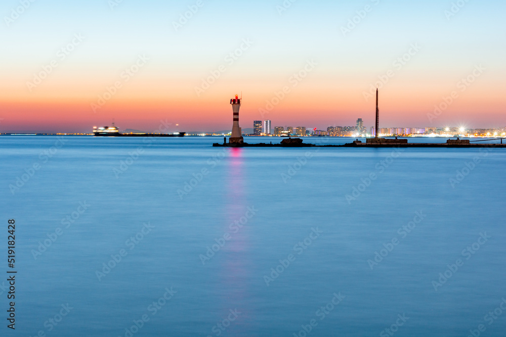 A landscape of sea with a lighthouse in the center and city lights in the foreground. At sunset.