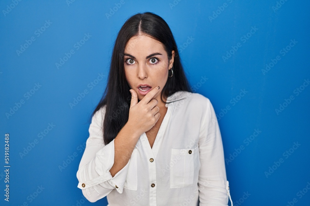 Young hispanic woman standing over blue background looking fascinated with disbelief, surprise and amazed expression with hands on chin Young hispanic woman standing over blue background looking fascinated with disbelief, surprise and amazed expression with hands on chin