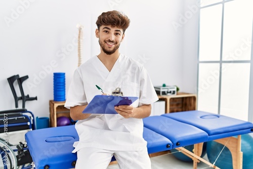 Young arab man wearing physiotherapist uniform writing on clipboard at clinic