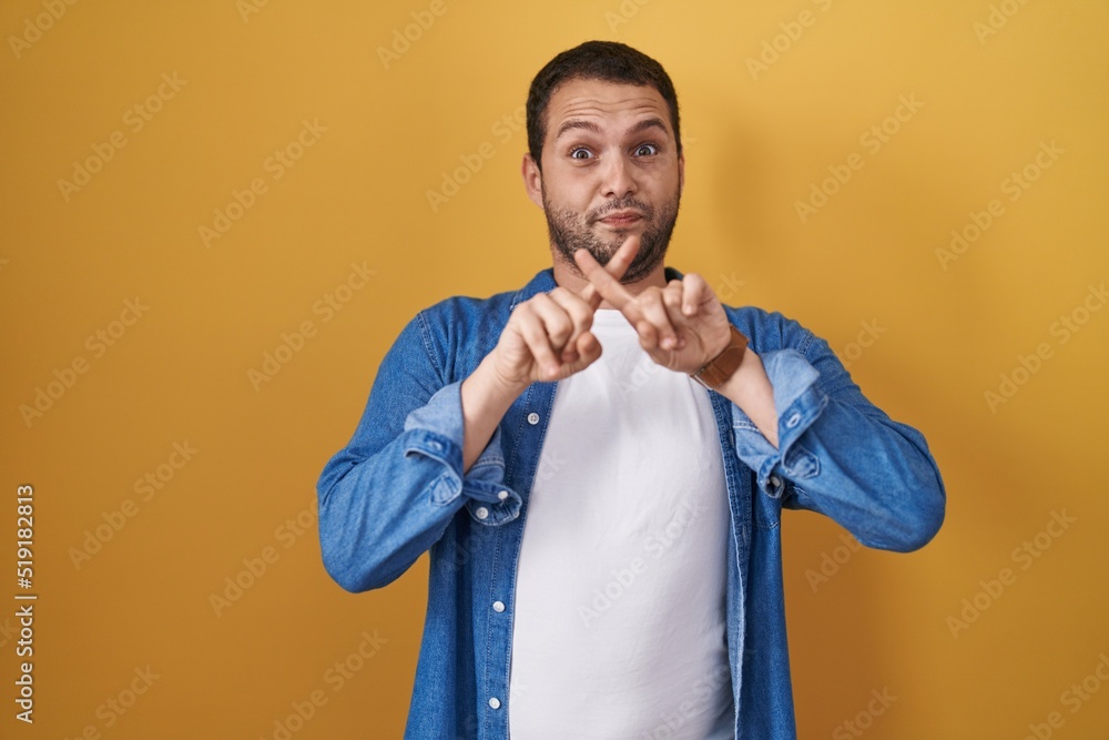 Hispanic man standing over yellow background rejection expression crossing fingers doing negative sign Hispanic man standing over yellow background rejection expression crossing fingers doing negative sign