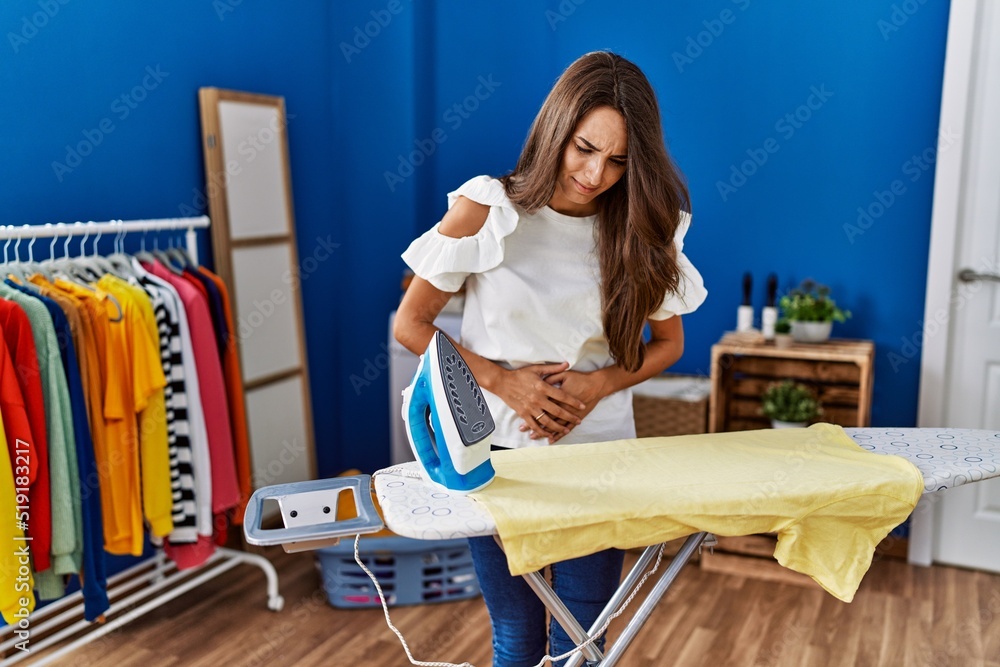 Young hispanic woman ironing clothes at laundry room with hand on stomach because nausea, painful disease feeling unwell. ache concept. Young hispanic woman ironing clothes at laundry room with hand on stomach because nausea, painful disease feeling unwell. ache concept.