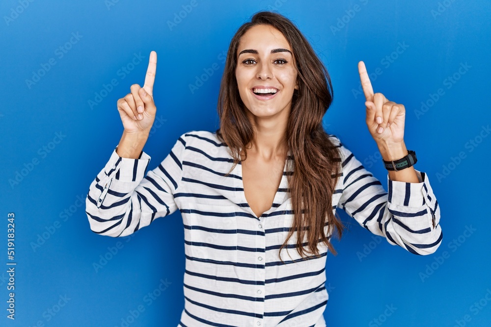 Young hispanic woman standing over blue isolated background smiling amazed and surprised and pointing up with fingers and raised arms. Young hispanic woman standing over blue isolated background smiling amazed and surprised and pointing up with fingers and raised arms.