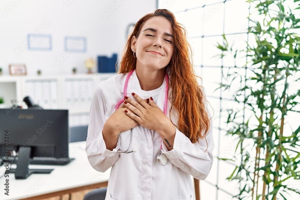 Young redhead woman wearing doctor uniform and stethoscope at the clinic smiling with hands on chest with closed eyes and grateful gesture on face. health concept. Young redhead woman wearing doctor uniform and stethoscope at the clinic smiling with hands on chest with closed eyes and grateful gesture on face. health concept.