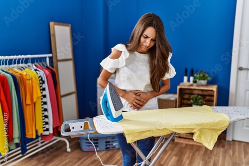 Young hispanic woman ironing clothes at laundry room with hand on stomach because nausea, painful disease feeling unwell. ache concept.
